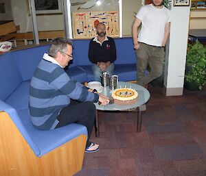 Male expeditioner cuts a cake, sitting on a circular lounge, cake on a low table.