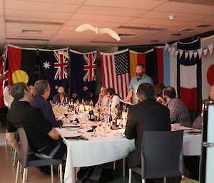 In the middle of a room lined with large flags, a table is set for dinner. Expeditioners sit around it, while a bearded male stands and reads a toast to New Zealand.