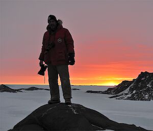 Kim, the station chef, stands on a rock at Colbeck. He is holding a camera, and the sun is rising brilliantly behind him.