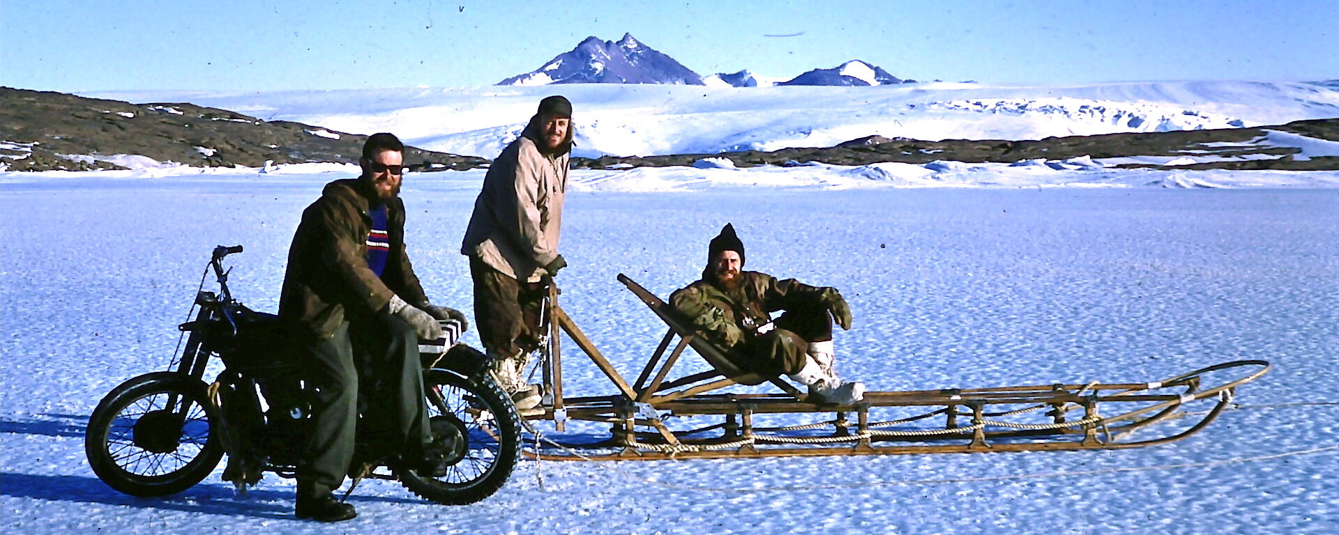 A Velocette motorcycle parked on the sea ice next to a dogsled and three men in 1960 near Mawson station.