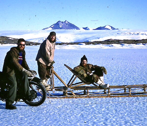A Velocette motorcycle parked on the sea ice next to a dogsled and three men in 1960 near Mawson station.