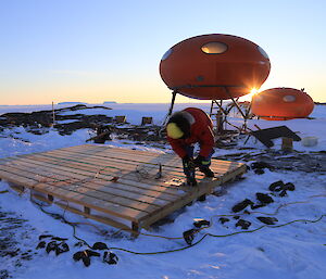 An expeditioner works on the wooden platform in front of the Melon hut, with a low sun on the horizon.