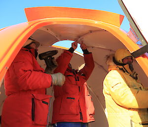 Three expeditioners are inside the partially constructed hut, filling the space, while they work on its construction