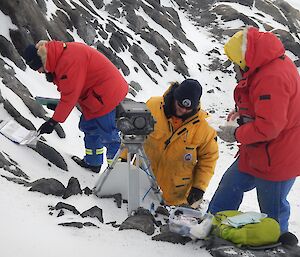 Three expeditioners work with and around a stationary penguin camera, with one behind ready to clean the lens.