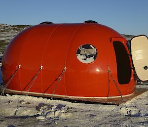 A small hut in the shape of a melon with a flat bottom, with door open and snow around