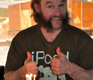 Male expeditioner with facial hair gives thumbs up in front of his cake which has lit candles on it.