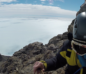 An expeditioner is climbing a rocky slope, their face covered by goggles, helmet and balaclava, and a cloud-like icy expanse below