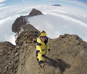 An expeditioner takes a selfie using a selfie stick and fish eye lens to show the top of a cliff, land and ice far below