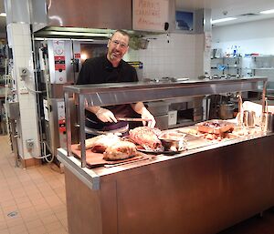 Tall male chef with facial hair behind bain-marie cutting a ham in a commercial kitchen — he smiles at the camera.