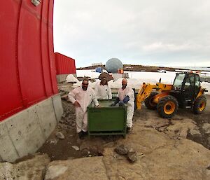 Three expeditioners in hazmat type suits stand outside around a large green box