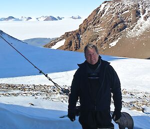 Station leader John Leben poses in front of a hut, not visible in photo. Snow and rocky slopes in background.