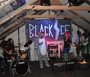 A band performs live inside at Mawson station with a large sign reading Black Ice in the background, the band’s name