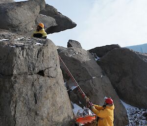 One man abseiling down a rock face with a stretcher and one man watching