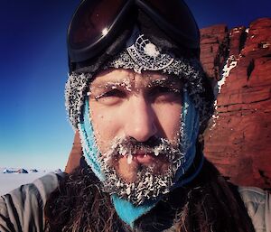 A man with a beard full of ice is taking a selfie, a tall rock visible in the background