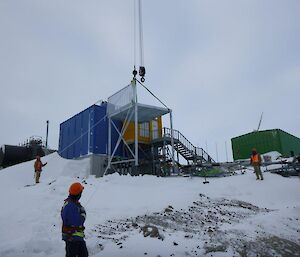 Several men use guide ropes to help lower a deck in place which sits at the end of a shipping container building