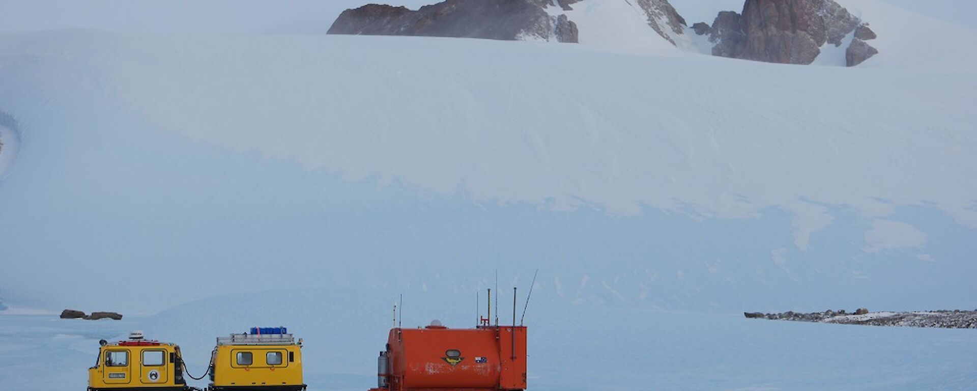 A yellow Hägglunds with a orange caravan attached to the back and a Mountain range in the distance