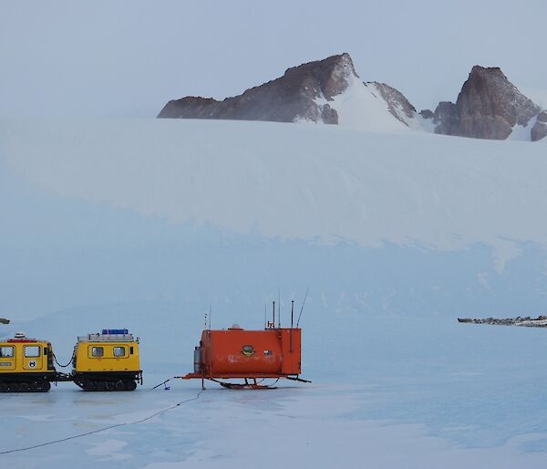 A yellow Hägglunds with a orange caravan attached to the back and a Mountain range in the distance