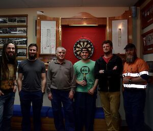 Six men posing in front of the darts board