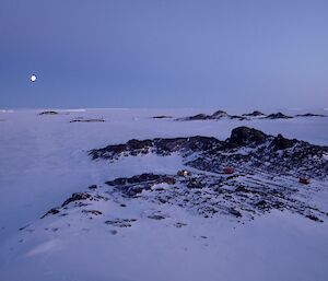 A small hut set amongst rocky hills and sea ice with a full moon