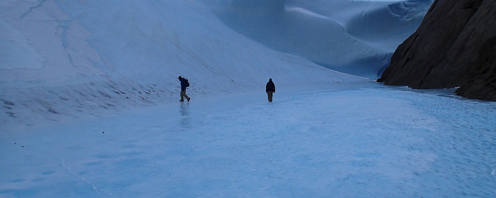 A large wave of ice at the bottom of a mountain with two expeditioners walking