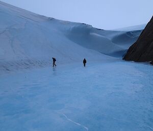 A large wave of ice at the bottom of a mountain with two expeditioners walking