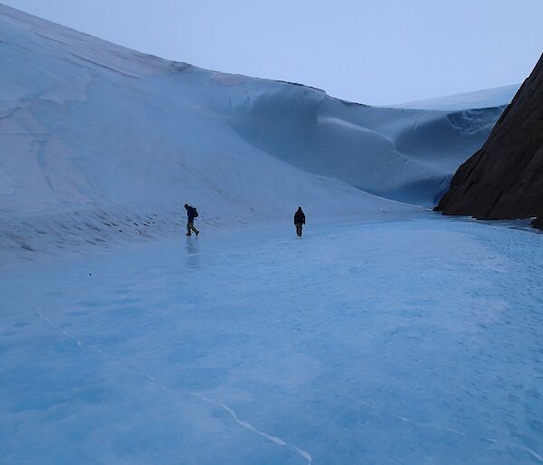 A large wave of ice at the bottom of a mountain with two expeditioners walking