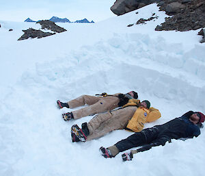 Three expeditioners lying in the snow to test the wind break they have dug out for their bivvies