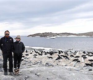 Two people standing near penguins with water and land behind them.