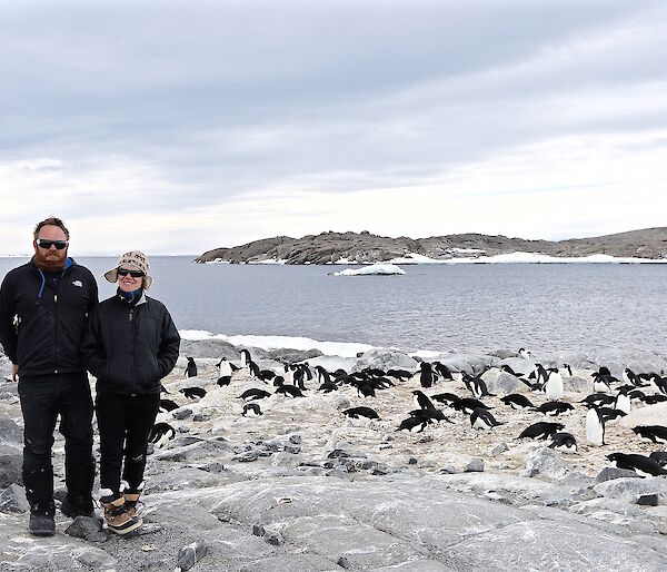 Two people standing near penguins with water and land behind them.