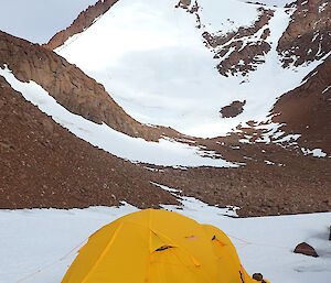 Yellow tent on the snow with rocks anchoring the sides at the base of Mt Hordern