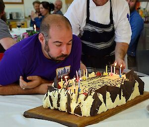 Leon is blowing out the candles on a large cream and chocolate covered cake