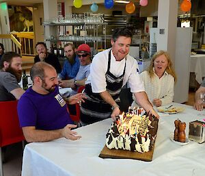 Benny is wearing a chef uniform and is presenting Leon with a large cream and chocolate cake