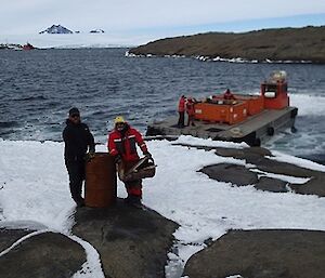 Two men are holding marine debris and old fuel drums with a barge and Mawson Station in the background