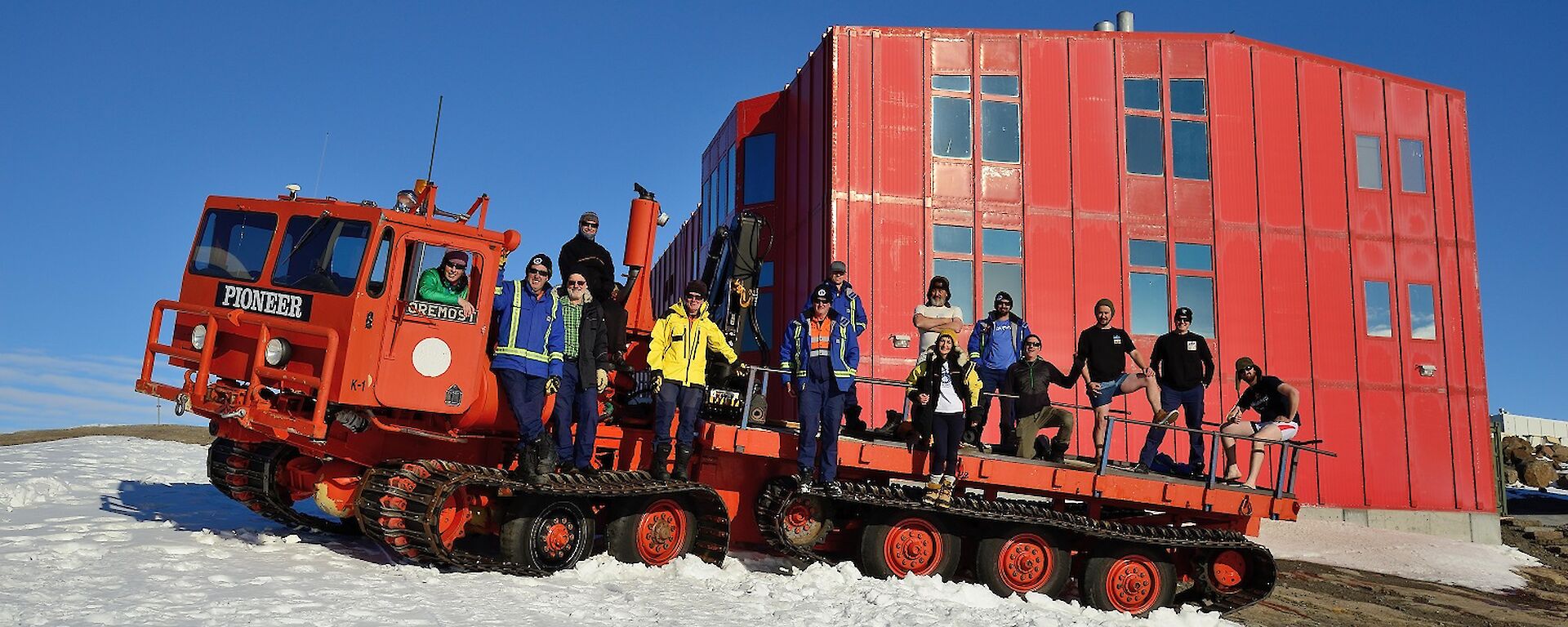 A large red over snow vehicle holds 14 expeditioners posing for a self timed photo in front of a large red shed and blue sky