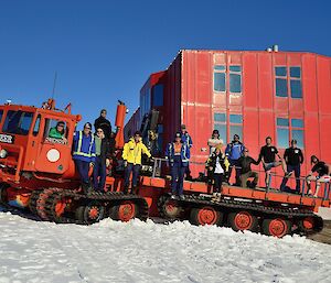 A large red over snow vehicle holds 14 expeditioners posing for a self timed photo in front of a large red shed and blue sky