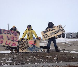 A group of 5 people are holding hand made signs that say “The boys are back in town”, “Back in 12 months”, “wrong way go back”.