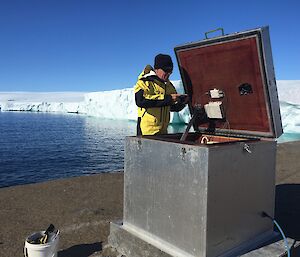 a man in a yellow jacket is inspecting an open metal box with a glacier ice cliff edge in the background