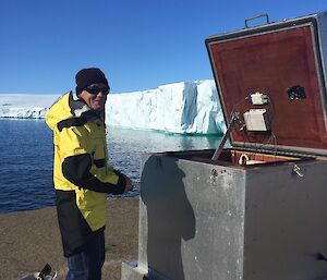 A man in a yellow suit stands in front of an open metal box with a glacier edge in the background