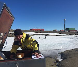 a man in a yellow jacket is crouching over a metal box with two Adélie penguins in the background