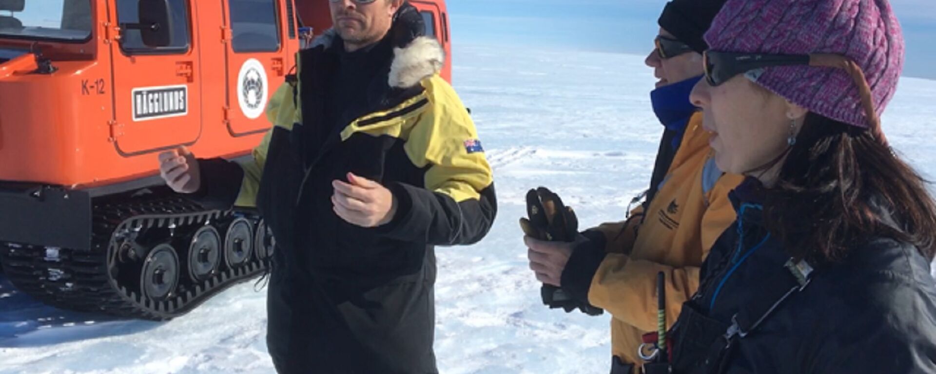 Three expeditioners stand on an icy plateau in front of an orange search and rescue equipped Hägglunds vehicle