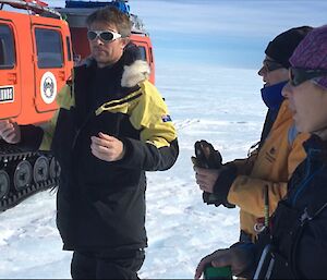 Three expeditioners stand on an icy plateau in front of an orange search and rescue equipped Hägglunds vehicle