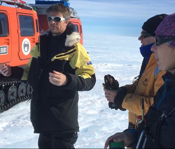 Three expeditioners stand on an icy plateau in front of an orange search and rescue equipped Hägglunds vehicle