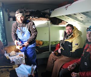 Three men and one woman wearing warm clothing are having a tea break in a small hut