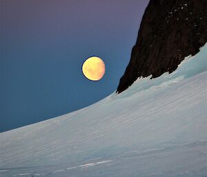 a three quarter yellow moon rises beside a mountain peak