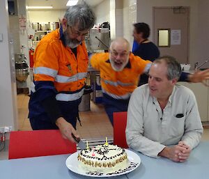 a man is leaping towards a birthday cake being cut by two men