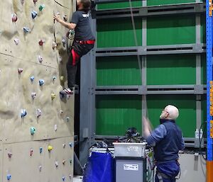 a man in a harness is climbing a rock climbing wall while another man holds a safety rope