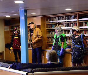 A man in a cap is talking to a group of people surrounded by three women in fancy dress costumes and masks