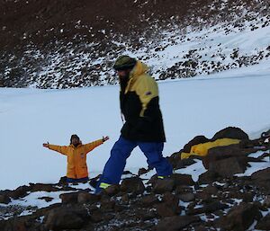 A man in a yellow coat holds his arms out to the side while anohter man in a yellow coat walks on rocks