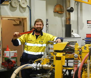 a man stands in front of a yellow machine holding a spanner over his shoulder