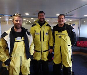 three men in yellow waterproof suits are standing in the lounge of a ship